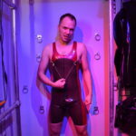 switchLDN in red and semi trans latex rubber wrestling suit in front of restraint wall pulling on clover nipple clamps