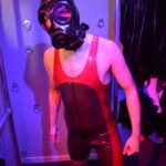 switchLDN in hooded s10 gas mask and red and semi trans latex rubber wrestling suit in front of restraint wall
